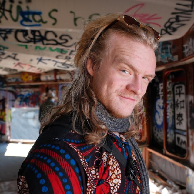 Bentley Perkins: shoulder-length fair hair with sunglasses pushed up into it, a grey knitted scarf, and a red and blue patterned shirt, standing in a room with graffiti-covered walls and ceiling.