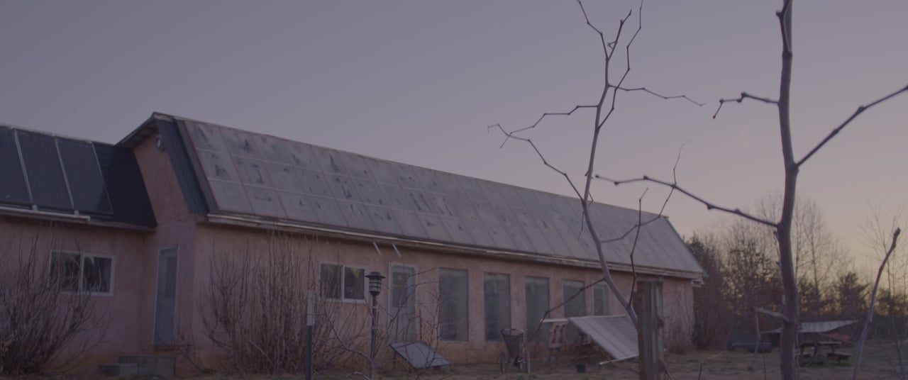 The main building at Living Energy Farm at dusk: a long earthen wall under a roof of solar panels, bare trees in front.